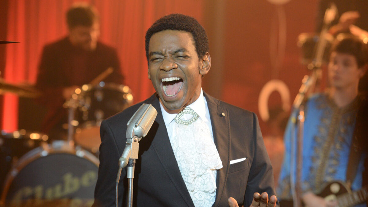 Fabrício Boliveira caracterizado como Wilson Simonal cantando no palco com banda ao fundo