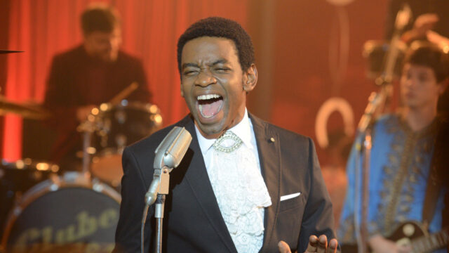 Fabrício Boliveira caracterizado como Wilson Simonal cantando no palco com banda ao fundo