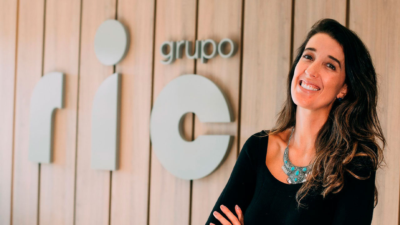 Laila Sol sorrindo com os braços cruzados em frente ao logo do Grupo Ric em parede de madeira