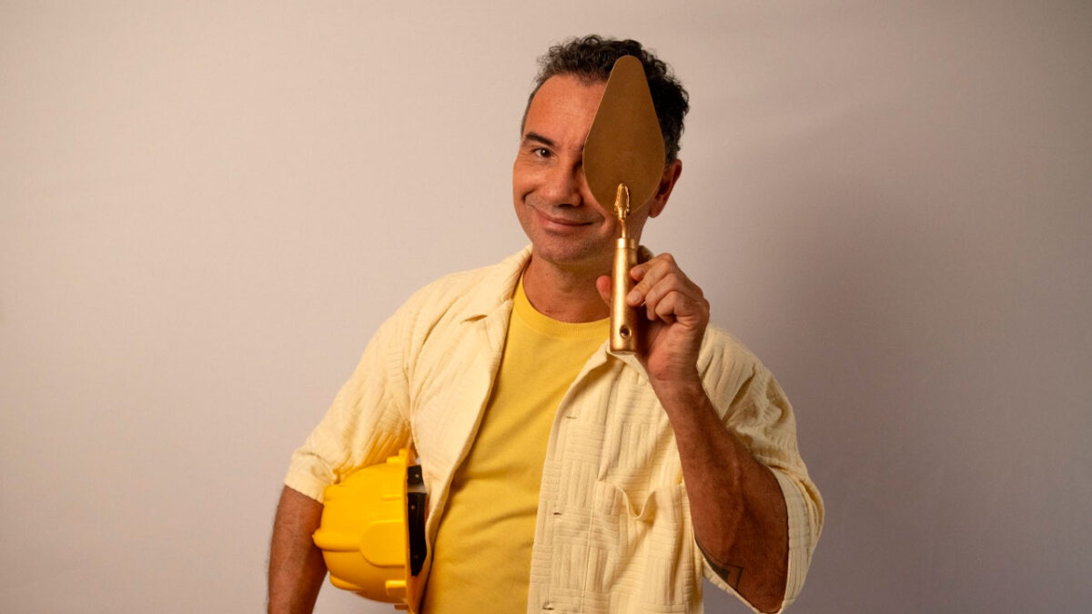 Marco Luque segura uma trolha dourada cobrindo um olho e capacete amarelo no braço em divulgação do Pedreiro Top Brasil