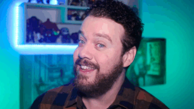Matheus Barros sorrindo durante gravação do programa Uplay com prateleira iluminada e objetos decorativos ao fundo