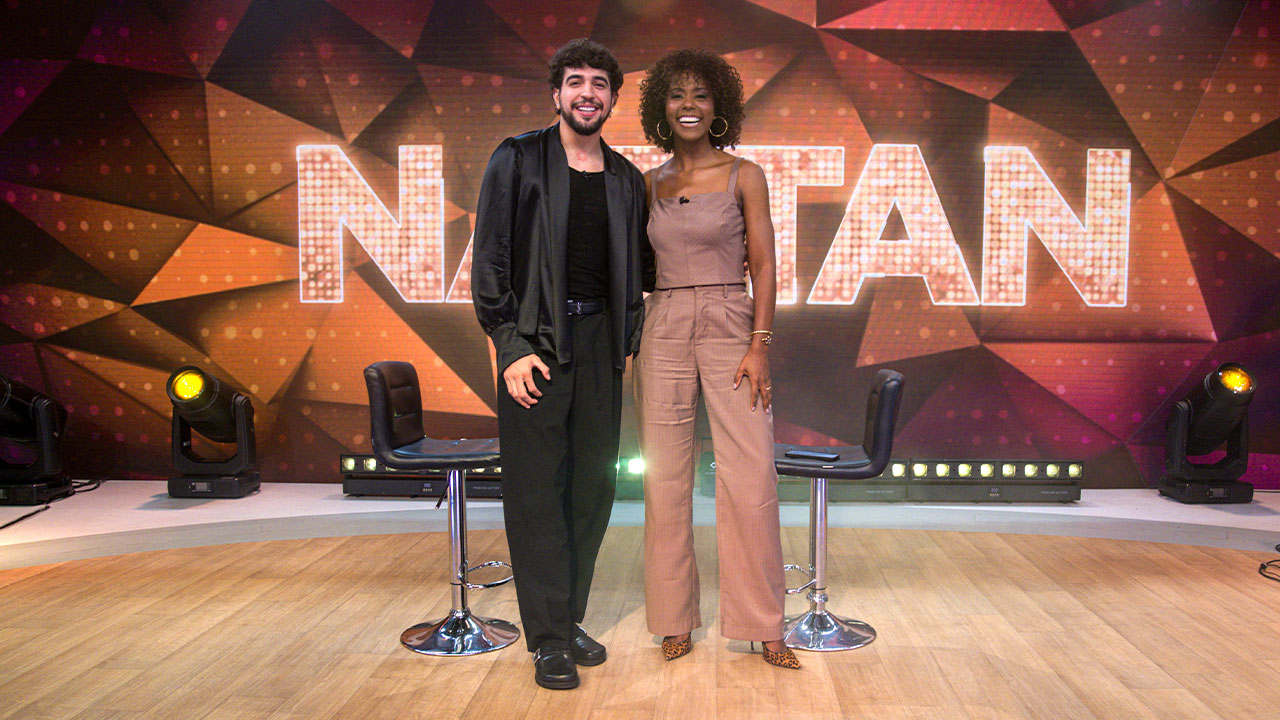 Nattan e Maju Coutinho posam sorrindo no palco do Fantástico diante do letreiro iluminado do programa