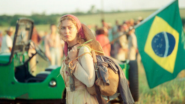 Patricia Pillar caracterizada como Luana carrega mochila em cena de O Rei do Gado com bandeira do Brasil ao fundo