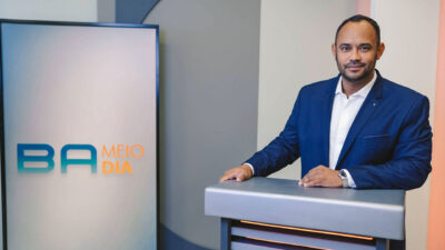 Vanderson Nascimento no estúdio do Bahia Meio Dia ao lado de painel com logotipo do telejornal