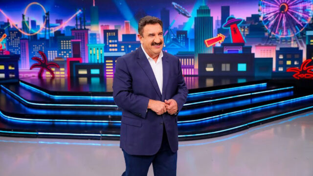 Carlos Massa, o Ratinho, de paletó azul no palco iluminado do Programa do Ratinho com cenário de cidade ao fundo