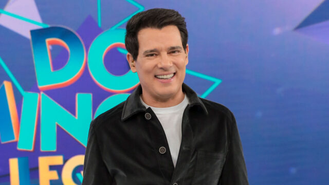 Celso Portiolli sorrindo no palco do Domingo Legal com o logotipo colorido do programa ao fundo