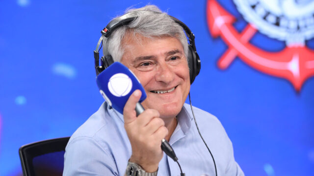 Cléber Machado com fone de ouvido segurando microfone com logo da Record em estúdio com fundo azul