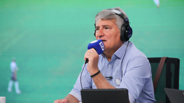 Cléber Machado com fone de ouvido e microfone da Record durante transmissão esportiva com campo ao fundo