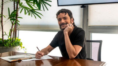 Fefito sentado à mesa sorrindo enquanto assina contrato com a TV Gazeta em ambiente interno iluminado