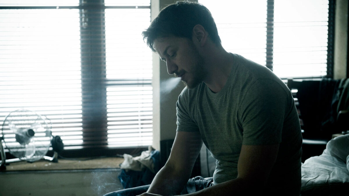 James McAvoy como Max Lewinsky sentado em quarto escuro soltando fumaça pela boca em Inimigos de Sangue