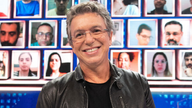 Boninho sorri em estúdio com painel de videochamadas exibindo rostos de várias pessoas ao fundo