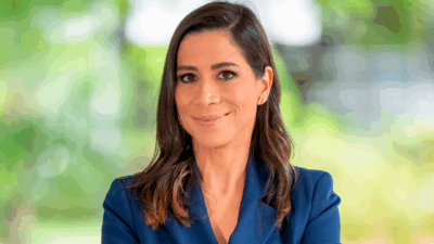 Julia Duailibi sorrindo com blazer azul marinho em cenário externo desfocado com vegetação ao fundo