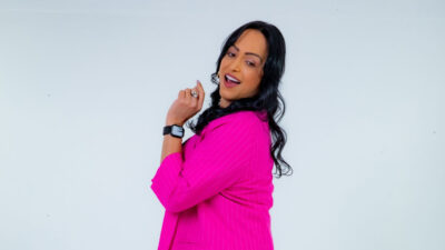 Lisa Gomes sorri de perfil usando blazer rosa e relógio digital diante de fundo branco