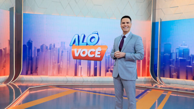 Luiz Bacci sorrindo no estúdio do Alô Você com o logotipo do programa ao fundo