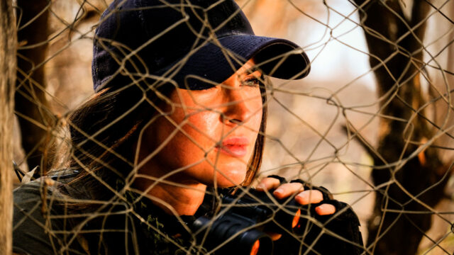 Samantha O'Hara (Megan Fox) observa atenta com binóculo atrás de rede camuflada em cena de Rogue (2020)