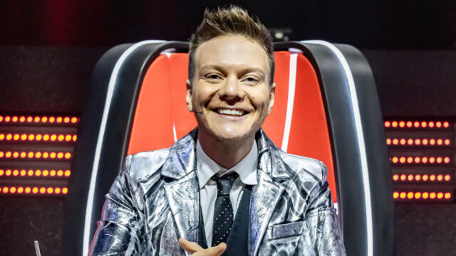Michel Teló sorri sentado na cadeira de técnico durante gravação do The Voice Brasil na Globo