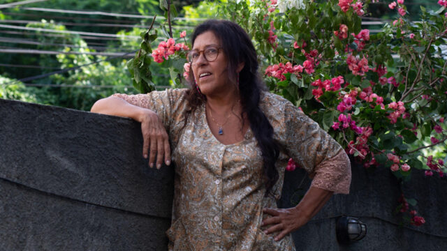 Regina Casé caracterizada como Lurdes apoia o braço em muro cercado por flores em Dona Lurdes - O Filme