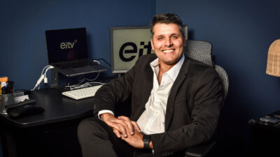 Rodrigo Cascão sorrindo sentado à mesa com computadores exibindo o logotipo da EiTV ao fundo