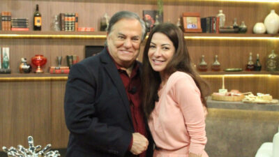 Ronnie Von e Ana Paula Padrão sorrindo lado a lado no cenário do programa Companhia Certa, com estante e mesa ao fundo