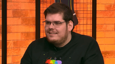 Casimiro Miguel sentado em estúdio com parede de tijolos laranja ao fundo usando camiseta preta com estampa colorida