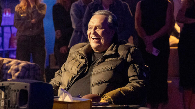 Dennis Carvalho sorrindo durante gravação do especial Tributo da TV Globo com pessoas ao fundo