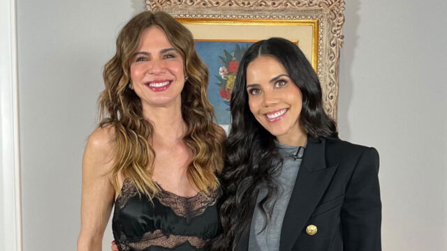 Luciana Gimenez e Daniela Albuquerque sorrindo juntas em frente a quadro decorativo em ambiente interno