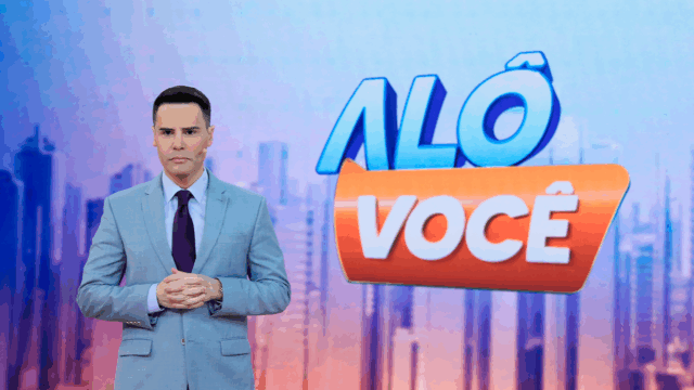 Luiz Bacci de terno claro diante do telão com o logotipo do Alô Você no estúdio do SBT