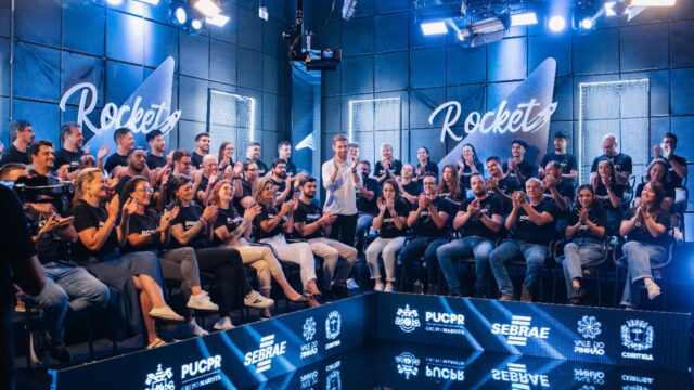 Participantes e apresentador do reality Rocket 2024 aplaudem em estúdio com logotipos da PUCPR e Sebrae ao fundo