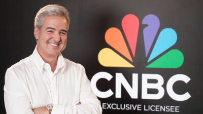 Thomaz Naves sorrindo com braços cruzados diante do logotipo da CNBC no anúncio do Times Brasil