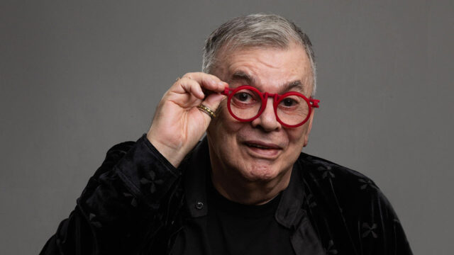 Walcyr Carrasco com óculos vermelhos redondos posa com a mão levantando a armação diante de fundo cinza