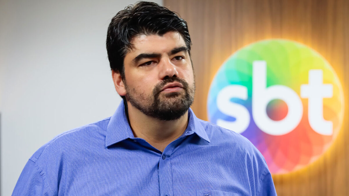 André Azeredo de camisa azul com expressão séria em frente ao logotipo do SBT