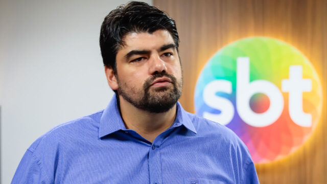 André Azeredo de camisa azul com expressão séria em frente ao logotipo do SBT