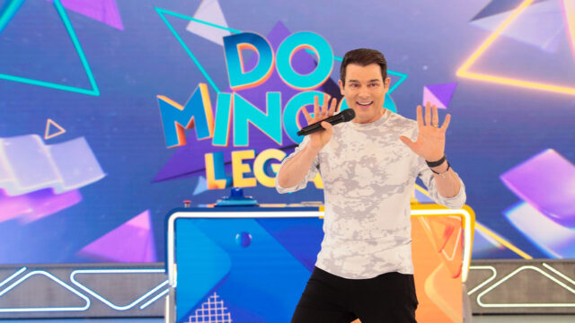 Celso Portiolli sorrindo com microfone na mão no palco colorido do Domingo Legal