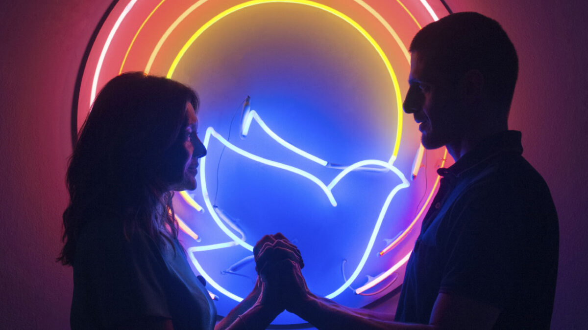 Joana (Dira Paes) e Danilo (Julio Machado) se olham e se tocam sob luzes neon coloridas em cena do filme Divino Amor (2019)