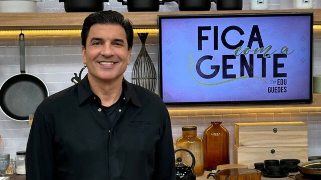 Edu Guedes sorrindo na cozinha do programa Fica com a Gente, exibido em tela ao fundo
