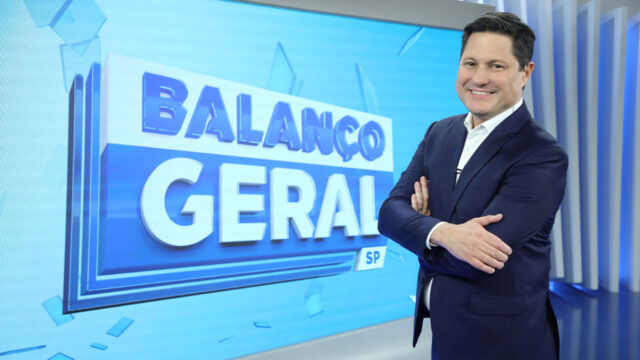 Eleandro Passaia sorrindo de braços cruzados ao lado do telão com logo do Balanço Geral SP