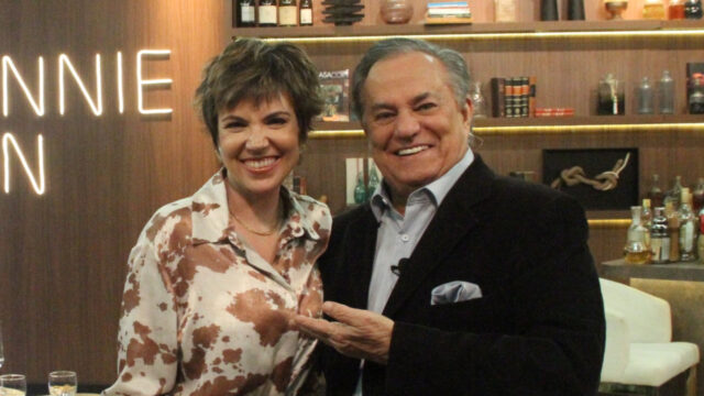 Glória Vanique e Ronnie Von sorrindo no cenário do programa Companhia Certa com estante de livros e garrafas ao fundo