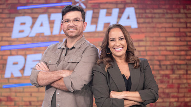 Guga Rocha e Renata Alves sorrindo lado a lado com braços cruzados no cenário do Batalha das Receitas do Hoje em Dia