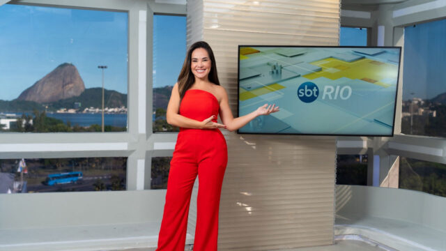 Isabele Benito sorrindo ao lado de tela do SBT Rio em estúdio com vista para o Pão de Açúcar ao fundo