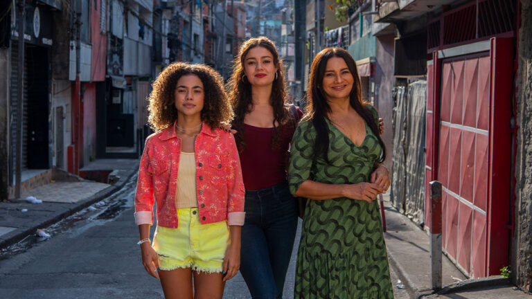 Joélly (Alana Cabral), Gerluce (Sophie Charlotte) e Lígia (Dira Paes) sorrindo em rua de comunidade na novela Três Graças