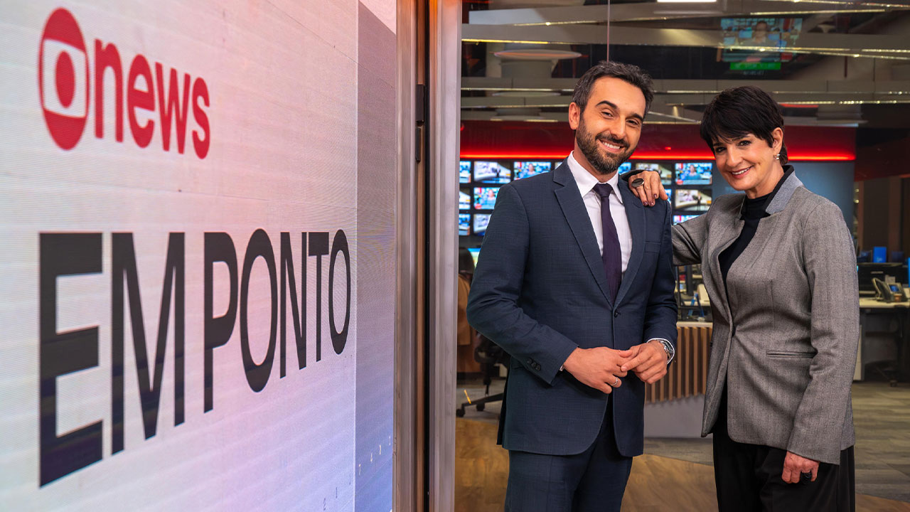 Victor Boyadjian e Mônica Waldvogel sorrindo ao lado do telão com o logo do Em Ponto na redação da GloboNews