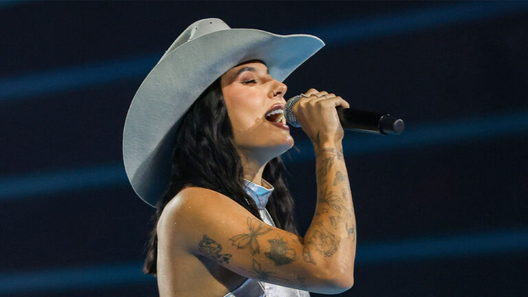 Ana Castela canta segurando microfone e usando chapéu branco em apresentação musical