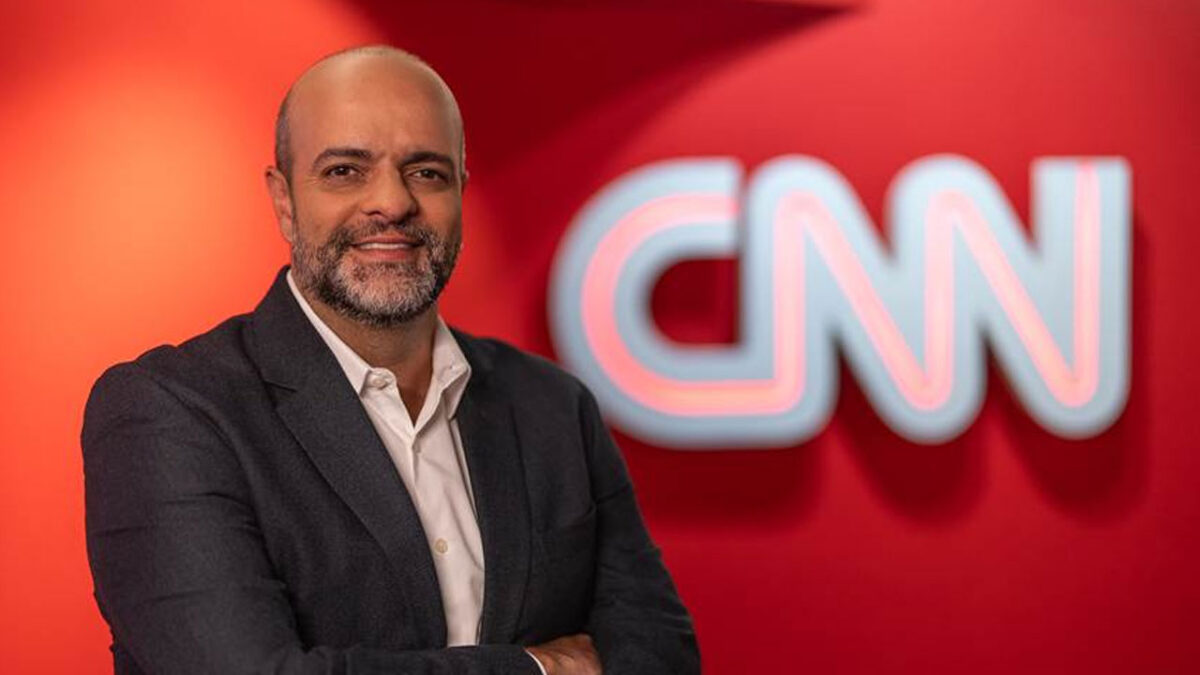 Bruno Bianchini de terno escuro e camisa clara posa sorridente em frente ao logotipo iluminado da CNN em fundo vermelho