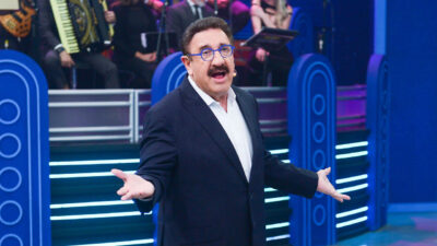 Carlos Massa o Ratinho de óculos azuis e terno escuro apresenta programa no palco com banda ao fundo