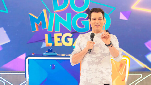 Celso Portiolli apresenta o Domingo Legal segurando microfone e levantando o dedo indicador em frente ao telão do programa no SBT