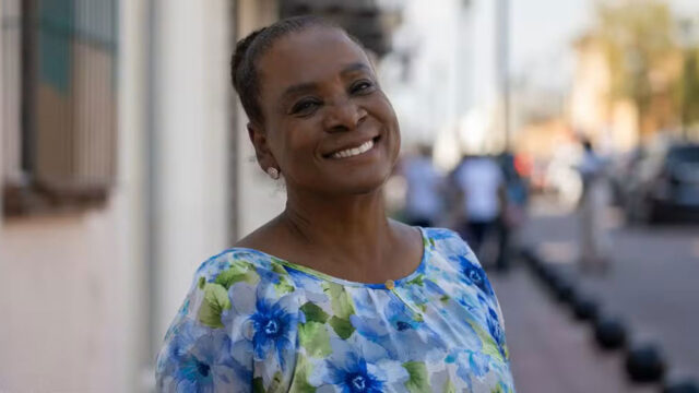 Dulcineia Novaes sorrindo com blusa estampada de flores azuis em rua ao ar livre