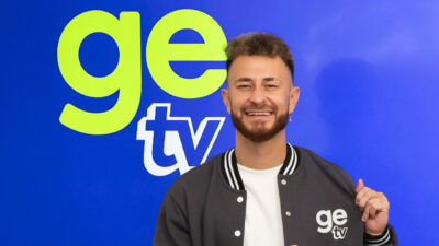 Fred Bruno sorridente veste jaqueta cinza da GE TV posando em frente ao logotipo do novo canal esportivo da Globo