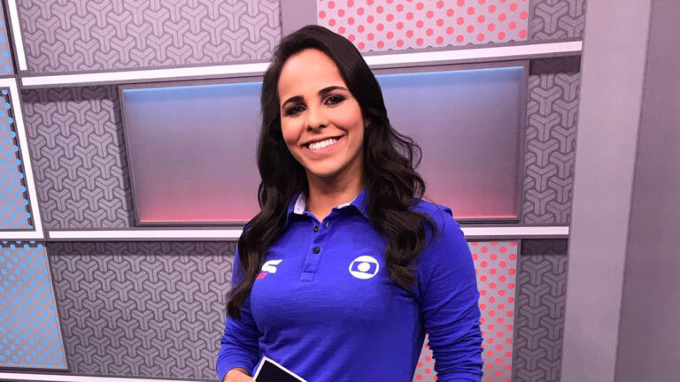 Isabelly Morais com camisa azul da Globo sorri no estúdio da emissora segurando tablet