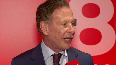 João Camargo de terno azul, camisa branca e gravata estampada falando em entrevista com microfone diante de painel vermelho