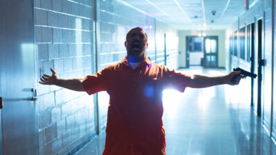 Mo Gallini como Ahmadi usando uniforme laranja de presidiário com arma em cena do filme Resposta Armada de 2017